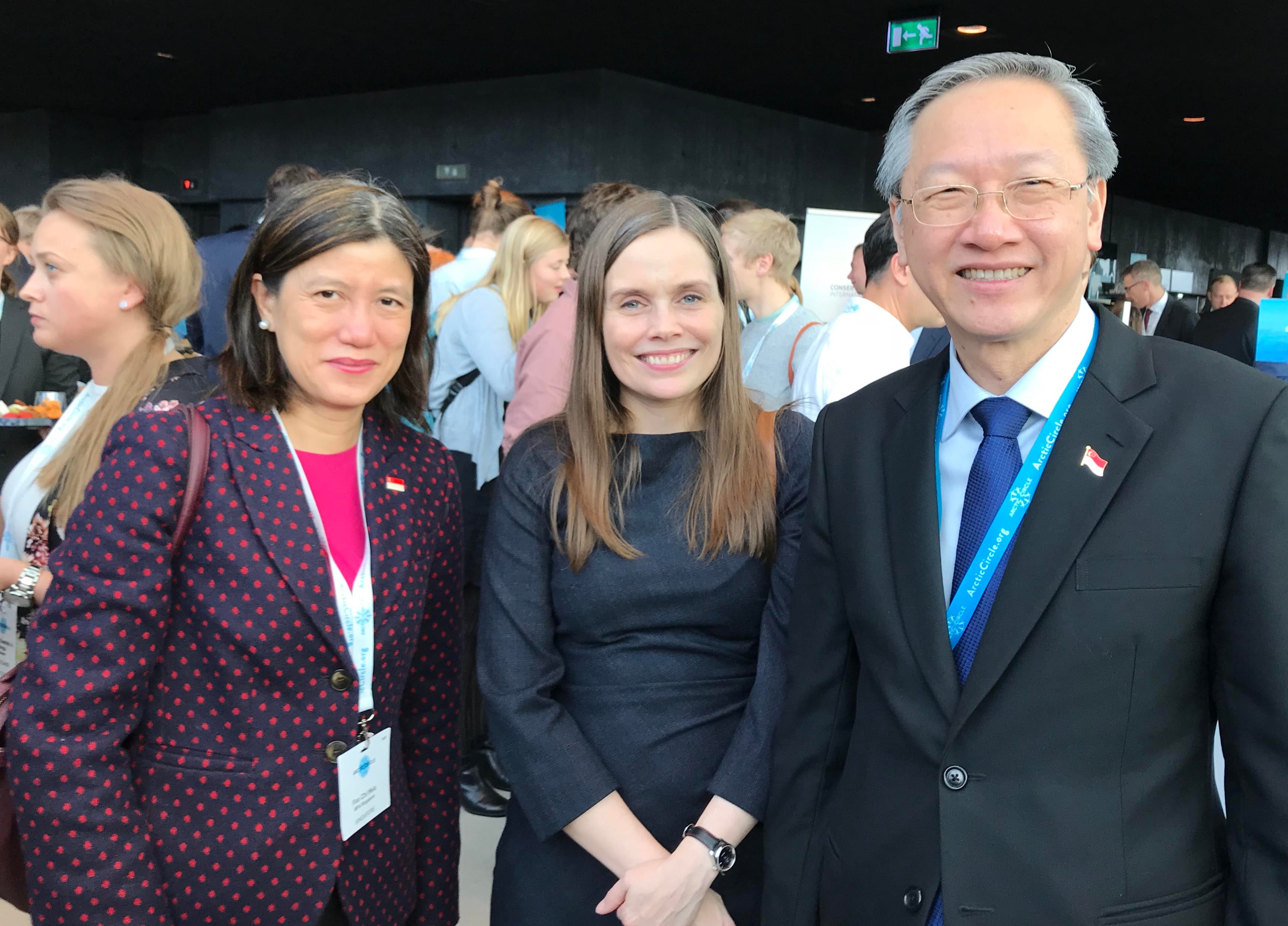 Three smiling people stand close, wearing business attire, badges. Crowd is visible in background.